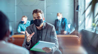 A man wearing a mask and holding a notepad speaks during a socially distanced meeting with a colleague.