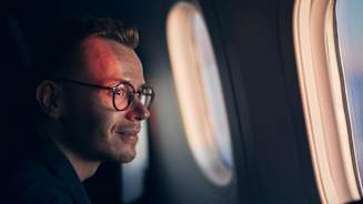 Man With Eyeglasses Traveling By Airplane. Passenger Looking Through Window During Flight At Sunset